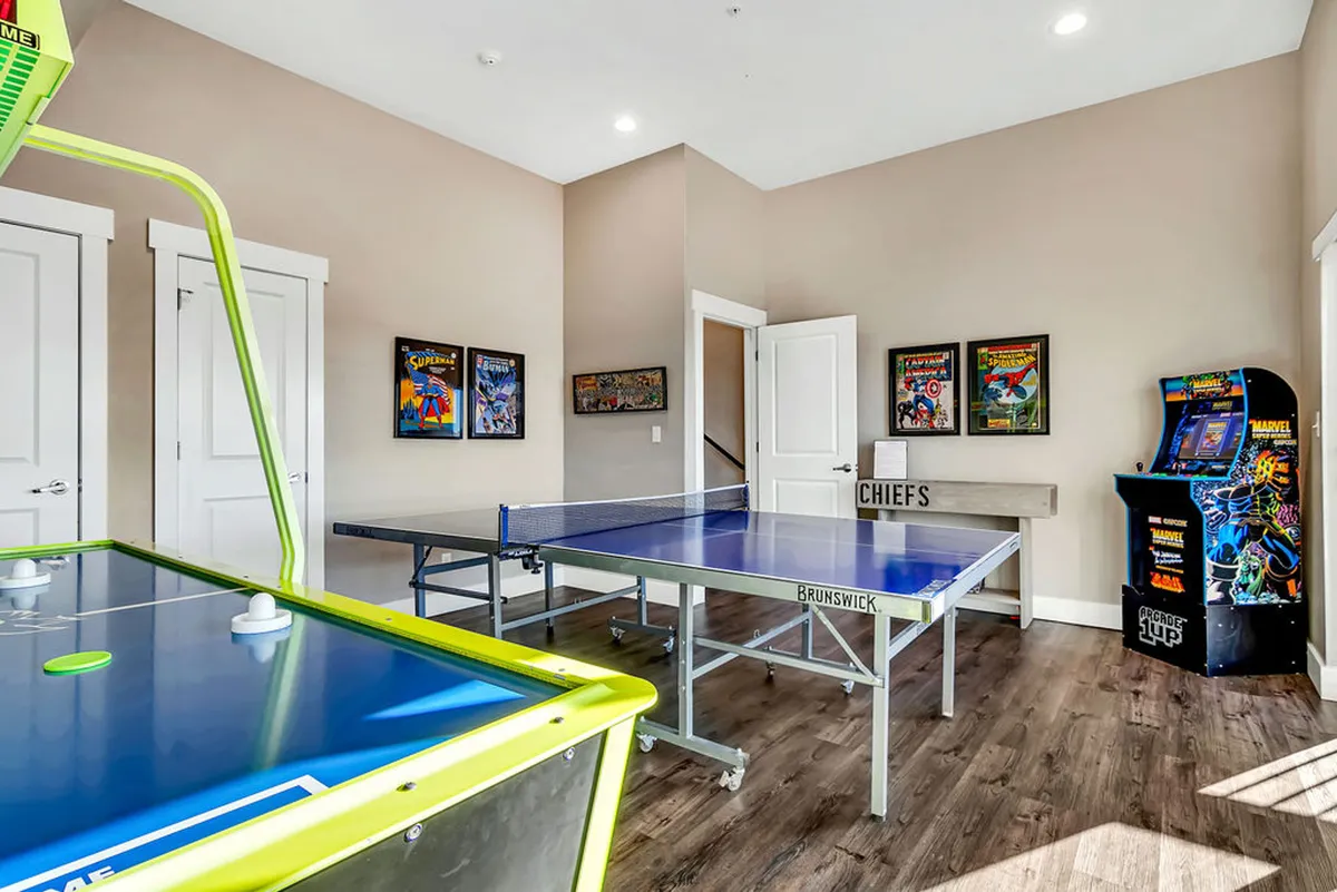 This interior image features an energetic game room with an air hockey table, ping pong table, and a bright arcade machine, all set against stylish decor and framed artwork. The space clearly welcomes social interaction, fun, and friendly competition.