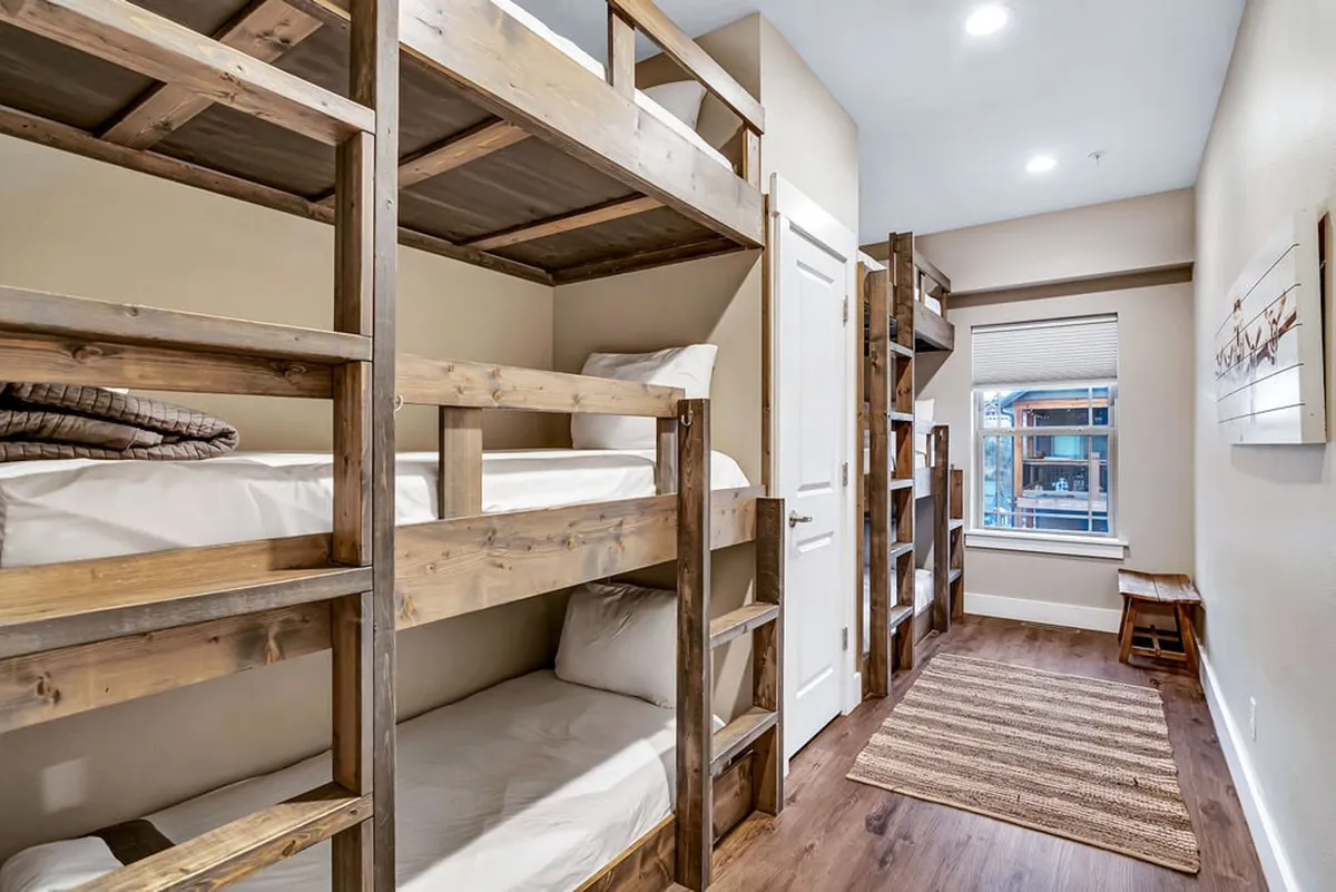 A bunkroom featuring sturdy wooden bunk beds, a sunlit window, wooden flooring, and a cozy rug. The space feels inviting and playful, with ample natural light and a comfortable corner for reading or playing, capturing the youthful and energetic spirit of the bunkrooms.