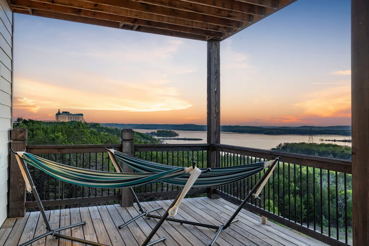 An exterior deck scene with hammocks swaying gently above a wooden deck, overlooking a golden sunset and tranquil lake view. The setup is inviting for outdoor relaxation and socializing.