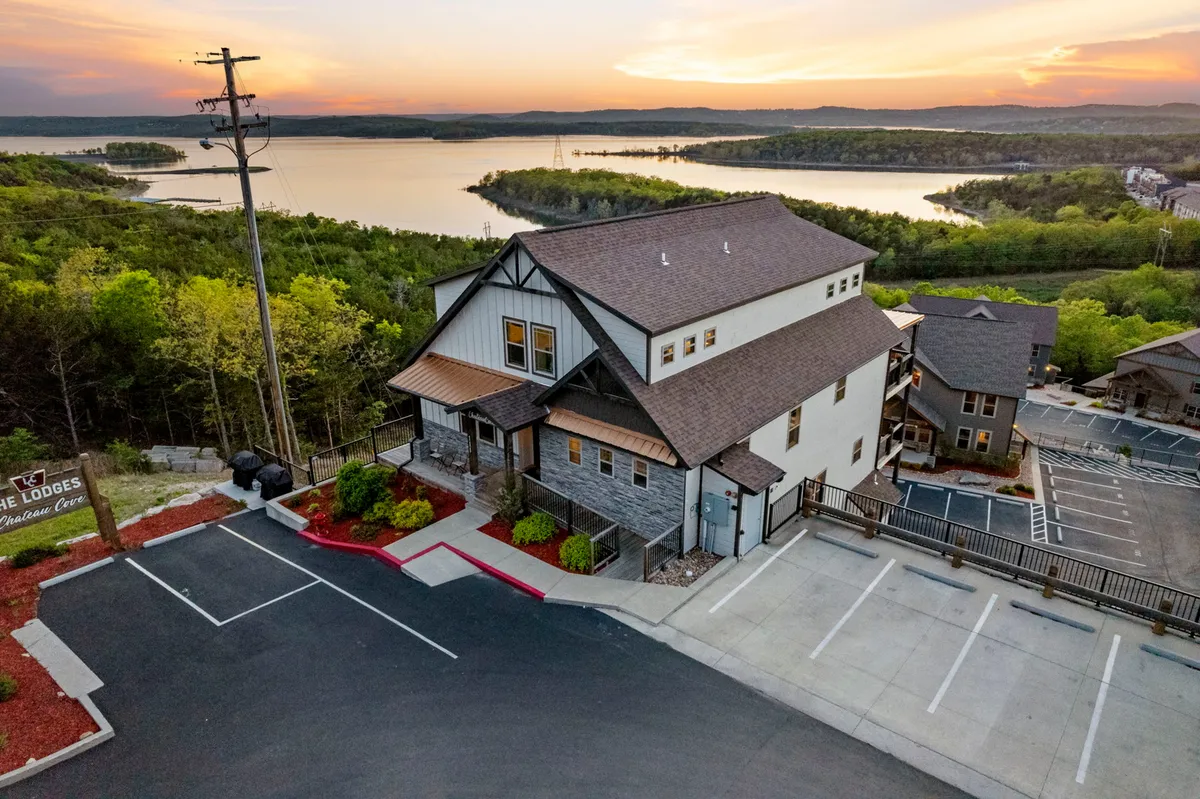 The exterior hero image showcases a grand, modern lodge nestled amid lush greenery, with expansive decks overlooking a tranquil lake and a stunning sunset sky. The serene water glimmers in the background, while the vibrant landscape and inviting architecture create an atmosphere of relaxation and togetherness.