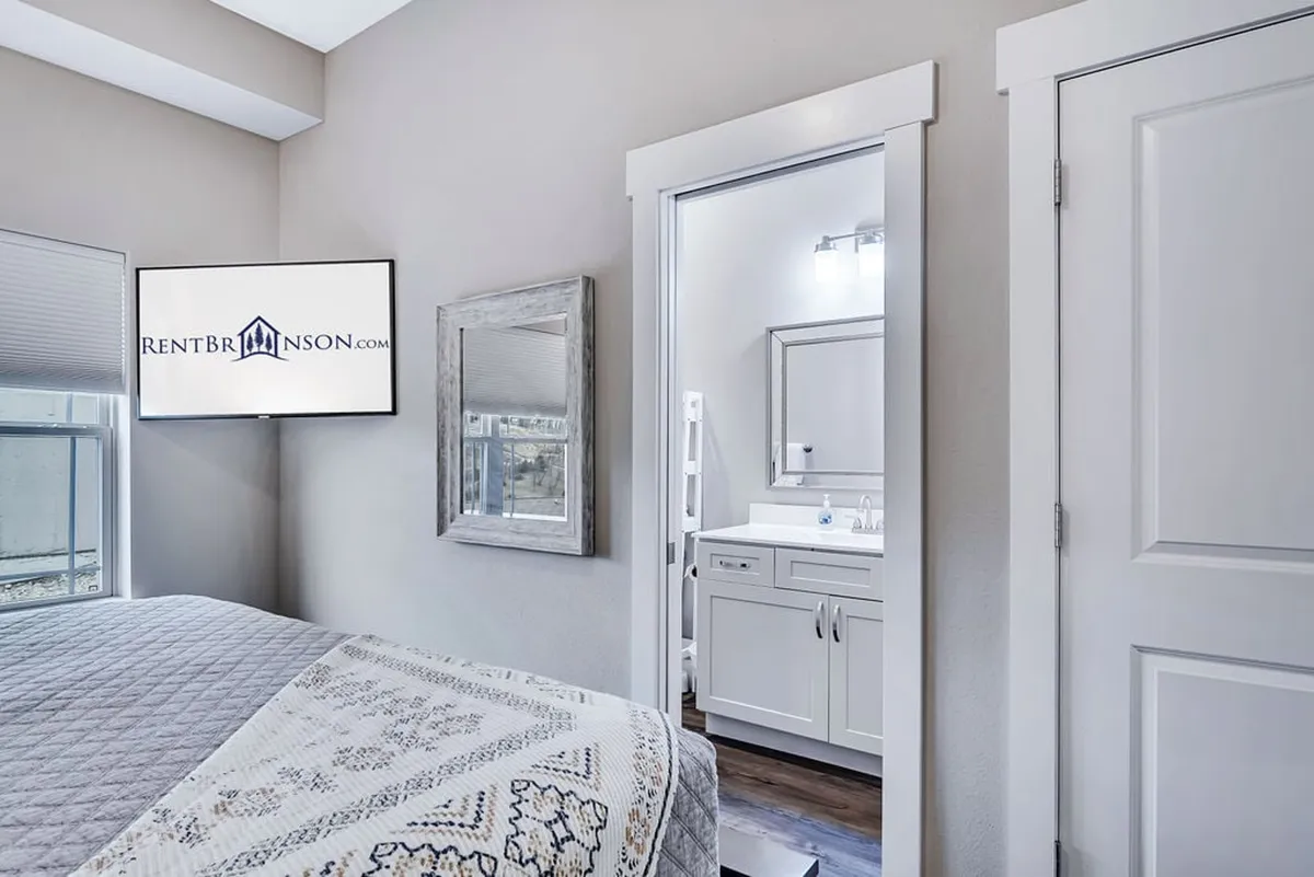 A bright bedroom with a bed adorned in patterned bedding, a wall-mounted smart TV, a mirror, and a glimpse of an ensuite bathroom entrance. The modern decor and layout suggest both relaxation and convenience, ideal for guests seeking in-room entertainment and privacy.