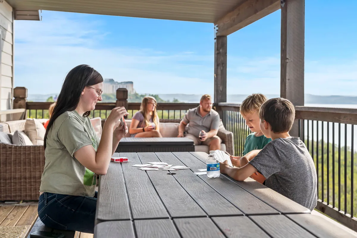 A lively scene of family members playing games around outdoor furniture, with a scenic landscape stretching into the distance. The joyful atmosphere underlines the fun, playful side of large gatherings, and the setting suggests ample space for group activities.